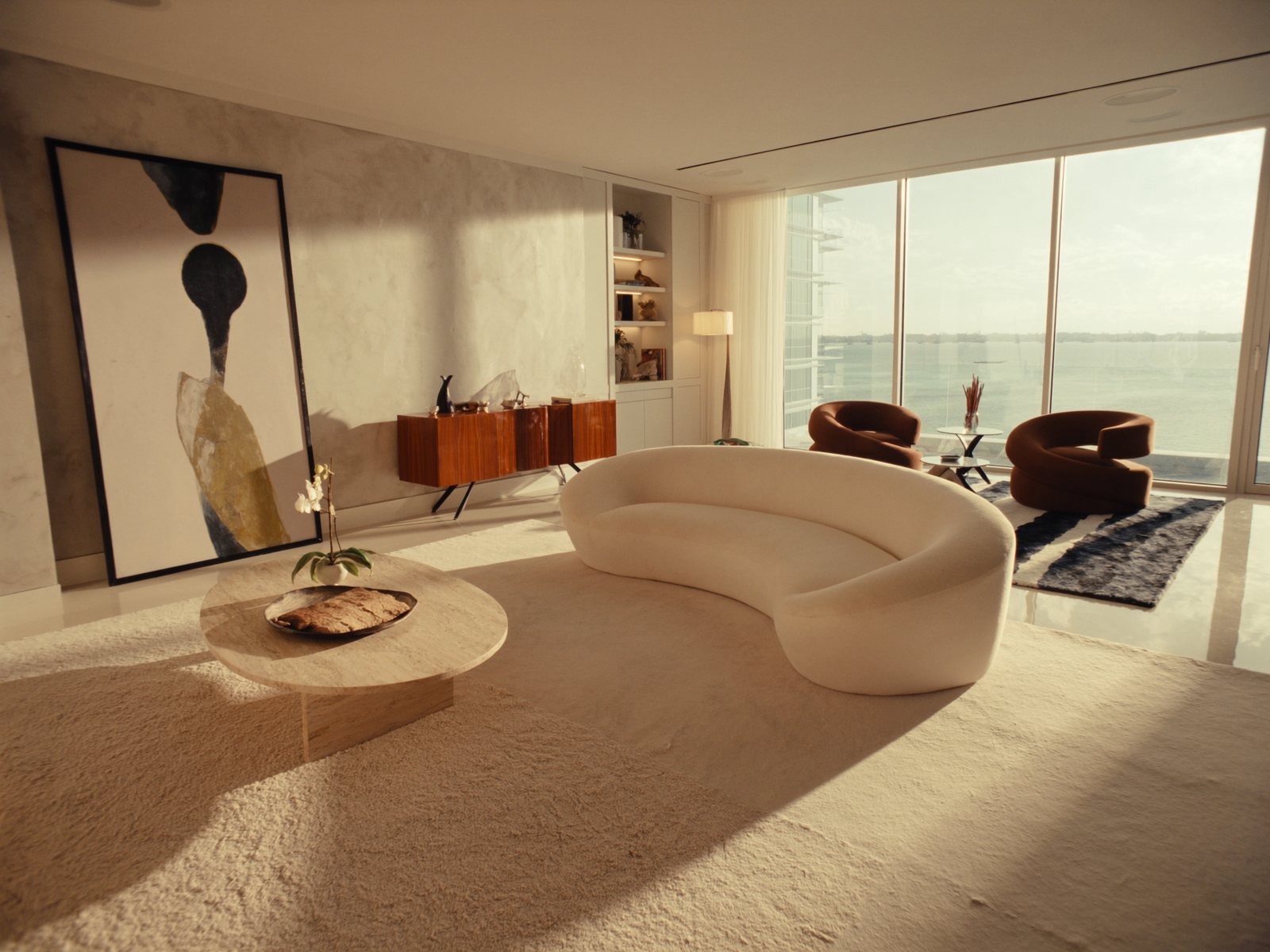 Luxury Miami condo living room with curved ivory bouclé sofa, cognac leather accent chairs, and travertine coffee table overlooking Biscayne Bay