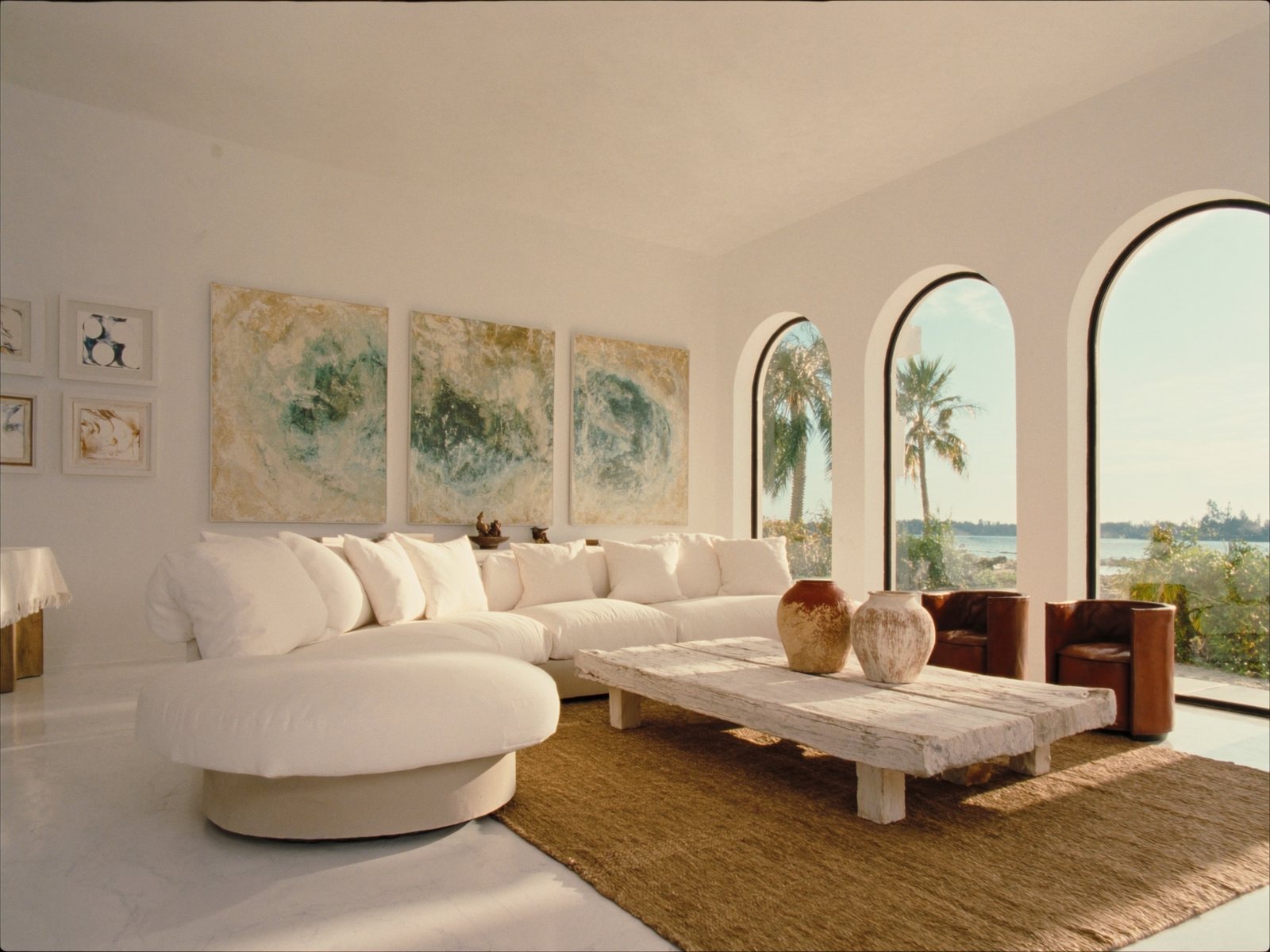 Sun-filled West Palm Beach estate living room with arched Mediterranean windows, curved ivory linen sectional, cognac leather accent chairs, and tropical garden views